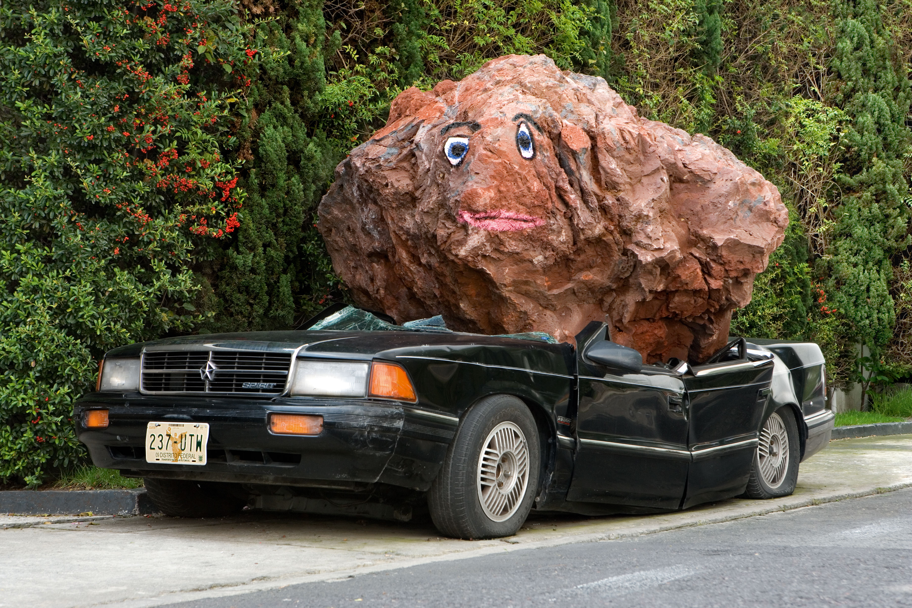 An old black car is squashed by a large brown blob, perhaps made with Papier-mâché, with eyes and a mouth