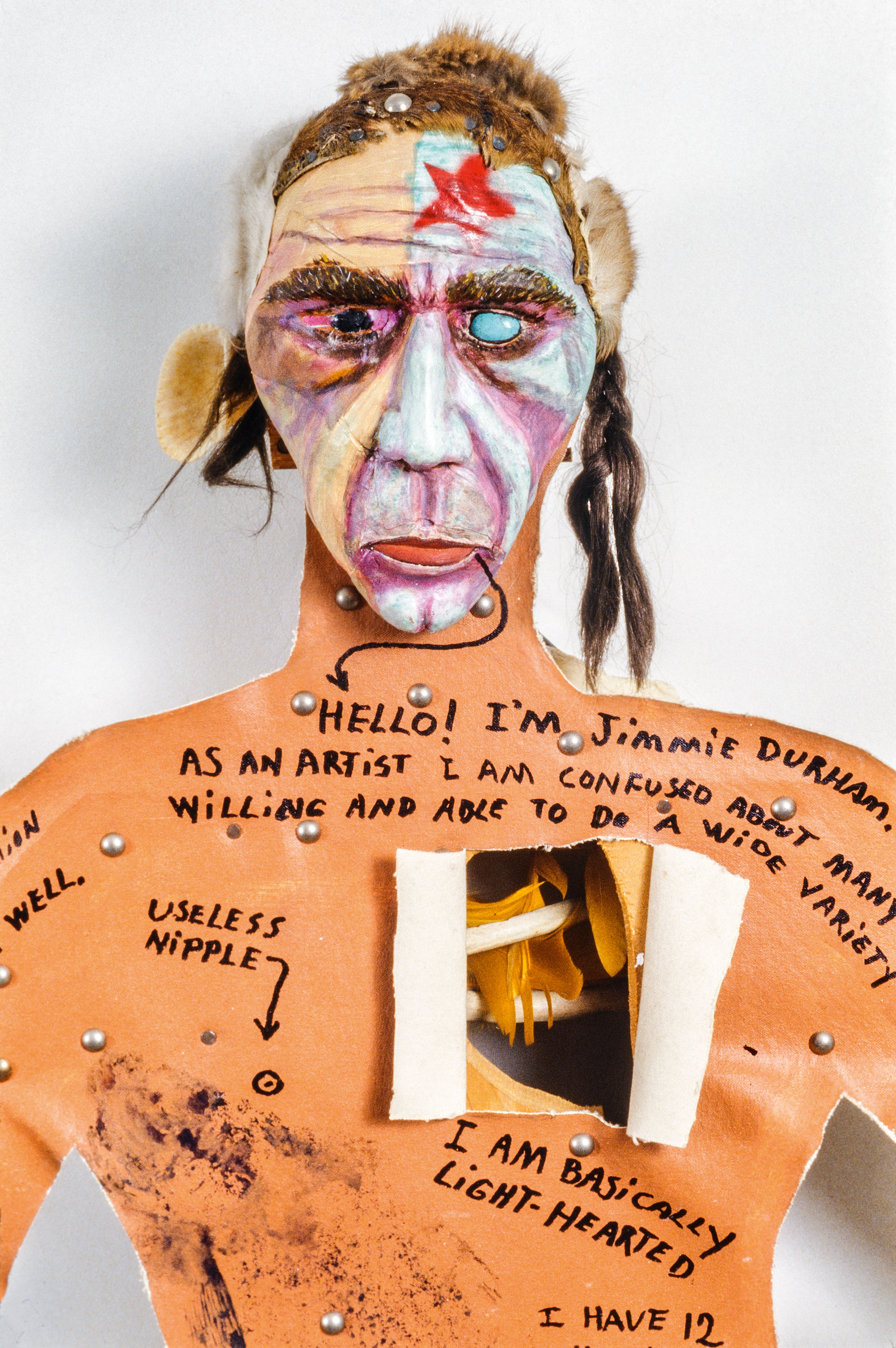 a composite sculpture of a man with a painted mask, hair made from fur and hair, and cardboard body with a hatch opened to show the heart cavity