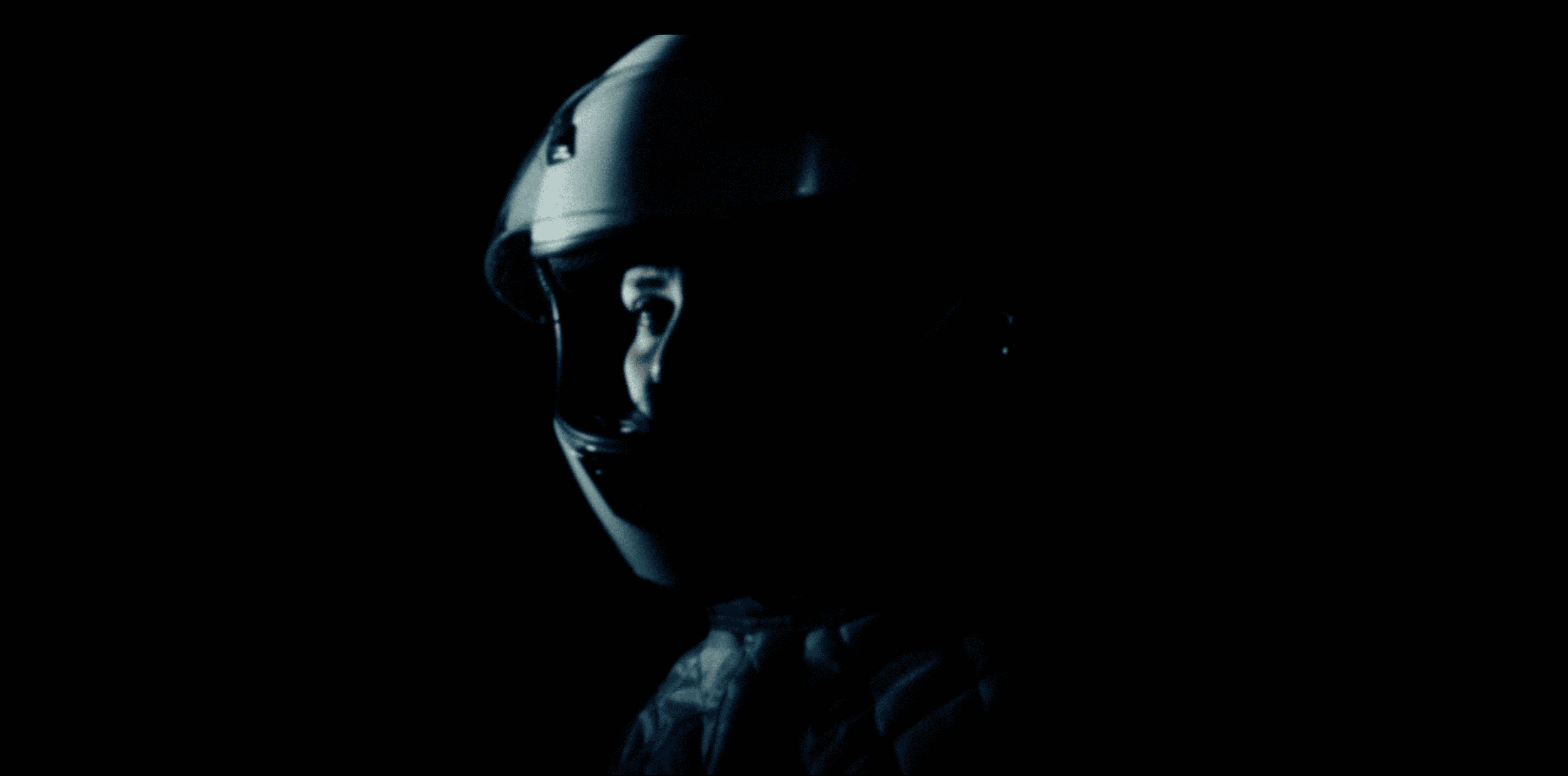 A woman in a racing driver's helmet looks at the camera. The background is dark.