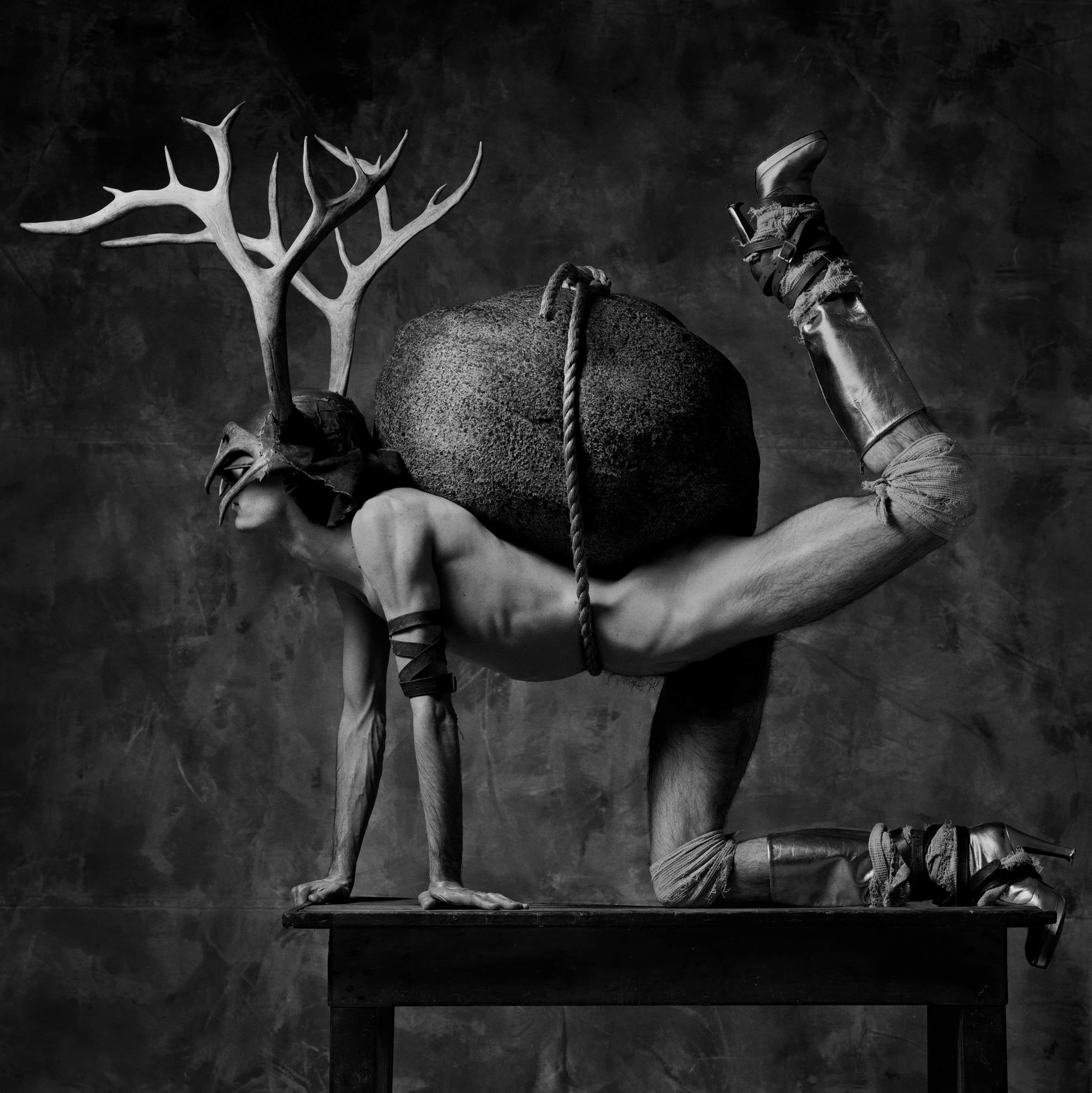 In black and white, a nude, young, slim yet muscular white man wearing large antlers poses side-on on all fours. He wear knee-high platform boots and his ankles are bound in leather strapping. Bandages swaddle his knees, and one leg is bent upwards off the ground. He balances on a table and a large rock is tied to his back with a rope. A smaller antler covers his face.