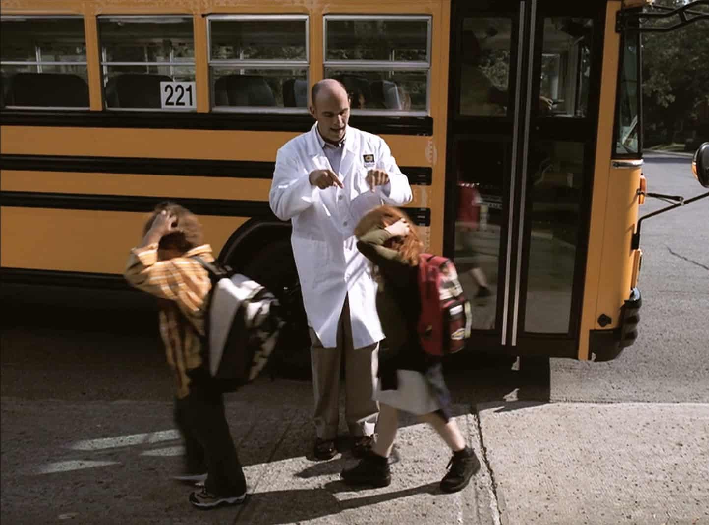 A pharmacist stands outside a school bus that has just dropped off children. He points to their heads because they have nits.